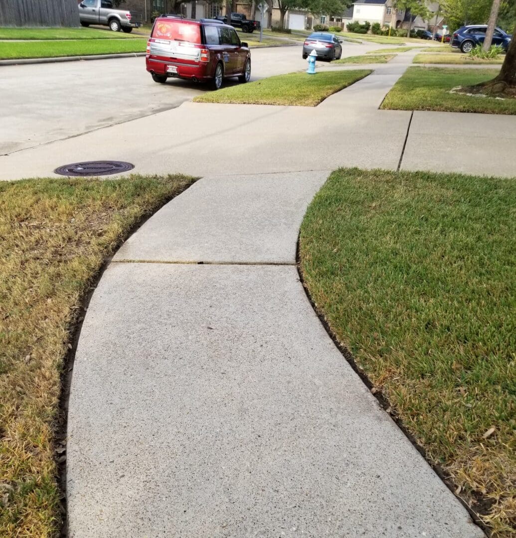 Curved concrete sidewalk bordered by grass and a street with parked cars.