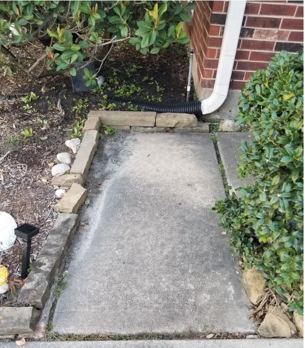 Concrete path with a section removed beside a brick wall and greenery.